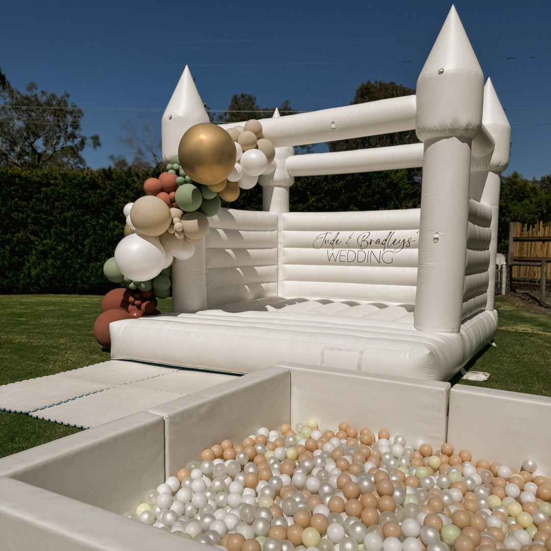 Alaska white bouncy castle with a gold, sage and rust balloon garland and Jude and Bradleys Wedding decal behind a ball pit