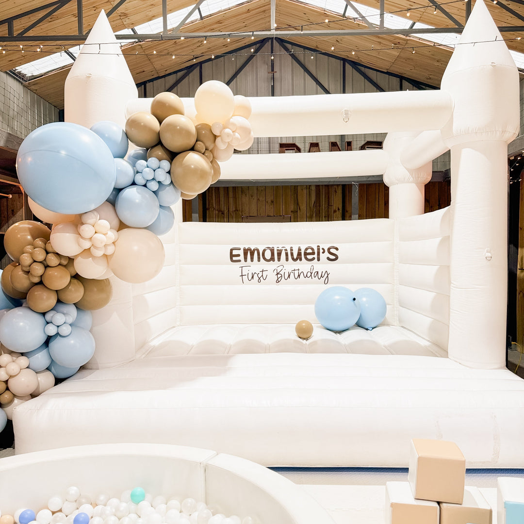 Alaska white bouncy castle in a warehouse venue with a blue, tan and cream balloon garland and Emanuel's First Birthday decal