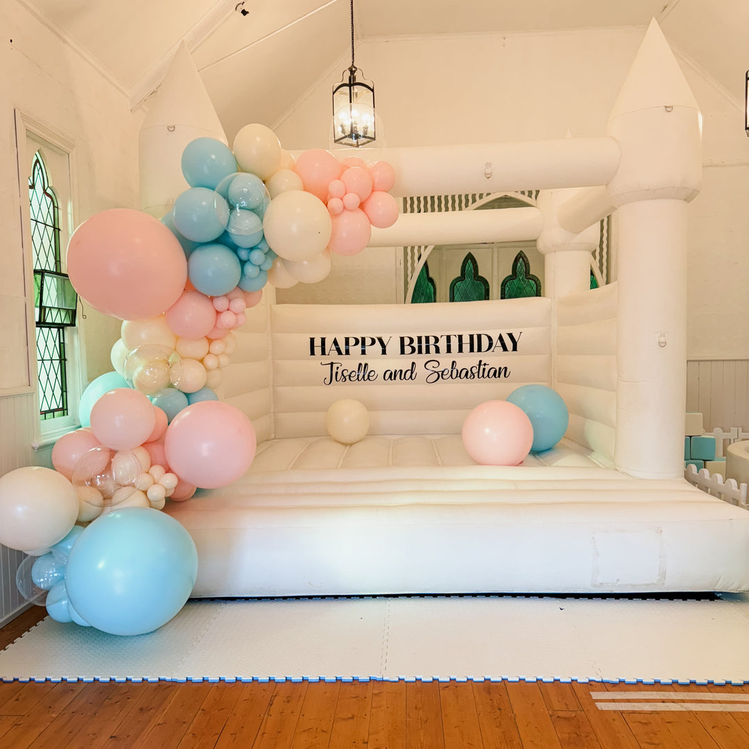 Alaska white bouncy castle in a chapel hall with a pastel pink and blue balloon garland and a Jiselle and Sebastian decal