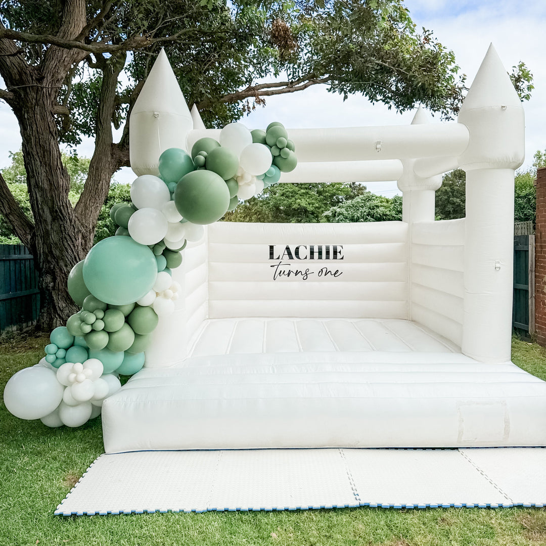 Sage and white balloon garland draped over the corner of a white bouncy castle with a Lachie turns one decal under a tree