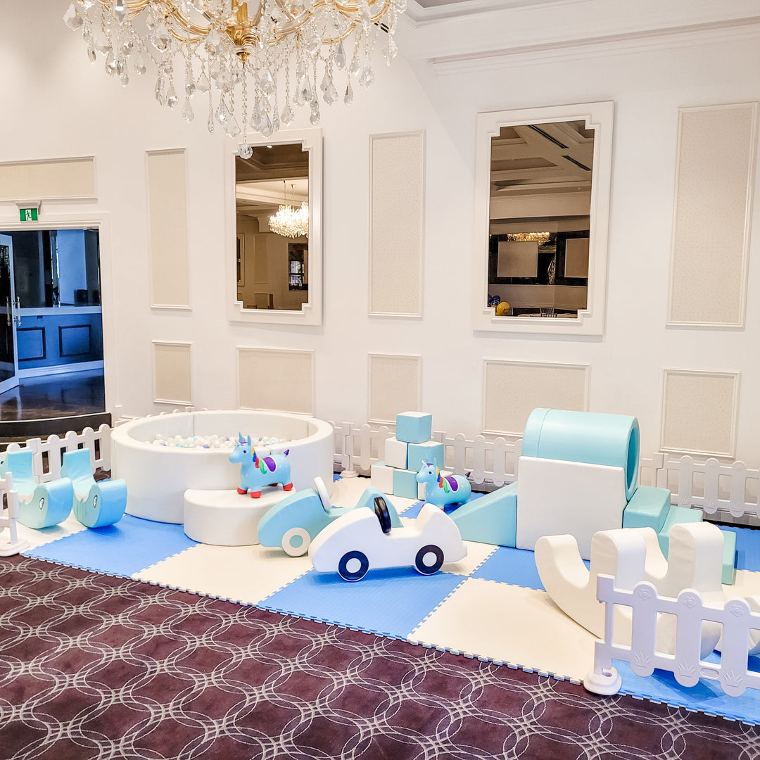 Baby blue dolphin rockers, ball pit, unicorn hoppers, foam cars and tunnel on a patterned ballroom carpet under a chandelier