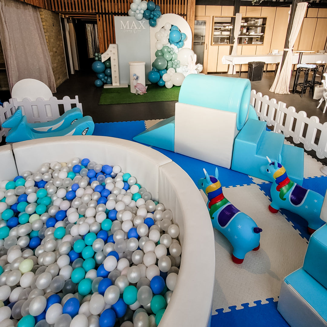 Close-up of the white ball pit filled with pearl, blue and aqua balls beside unicorn hoppers with a Max turns One arch behind