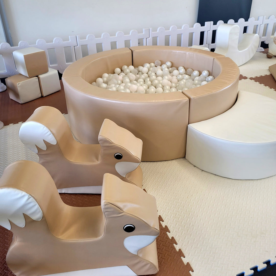 Close-up of two tan fox rockers in front of the tan ball pit of pearl balls, with foam blocks and a white half-moon step behind