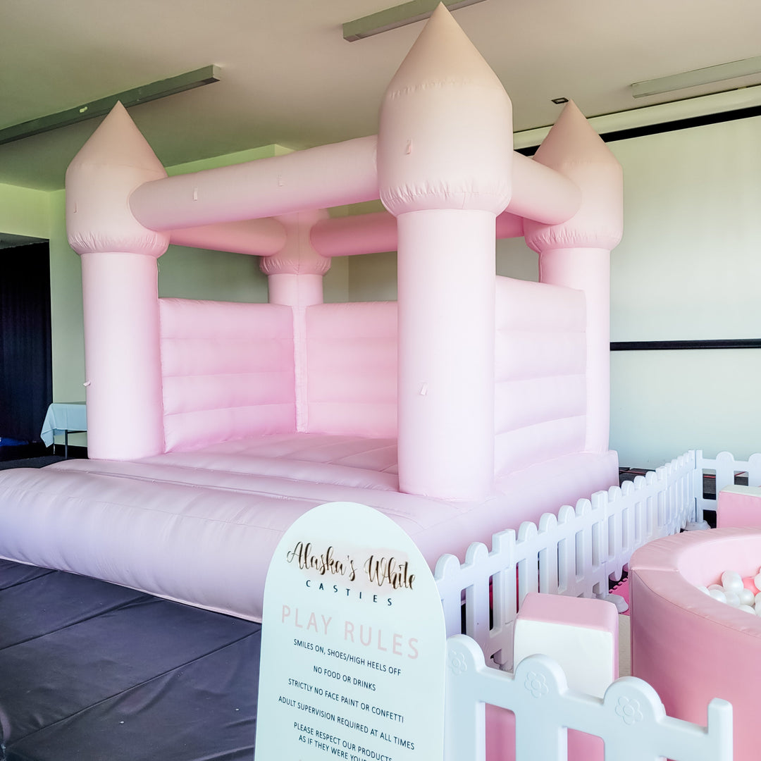Pink bouncy castle in a function room beside a pink ball pit, white picket fence and Alaska's White Castles play rules sign