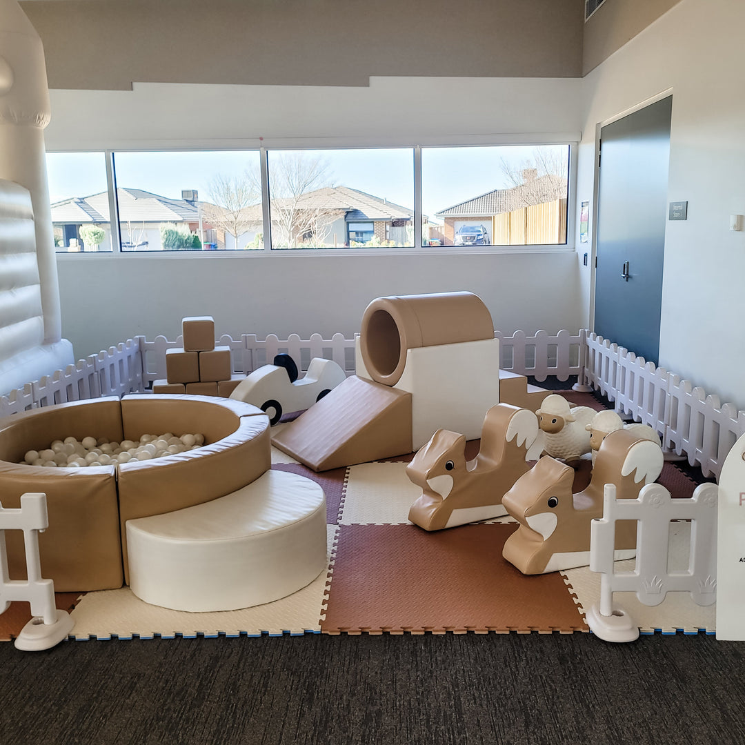 Tan soft play set with ball pit, tunnel slide, blocks, fox rockers and sheep hoppers on brown and cream mats by large windows