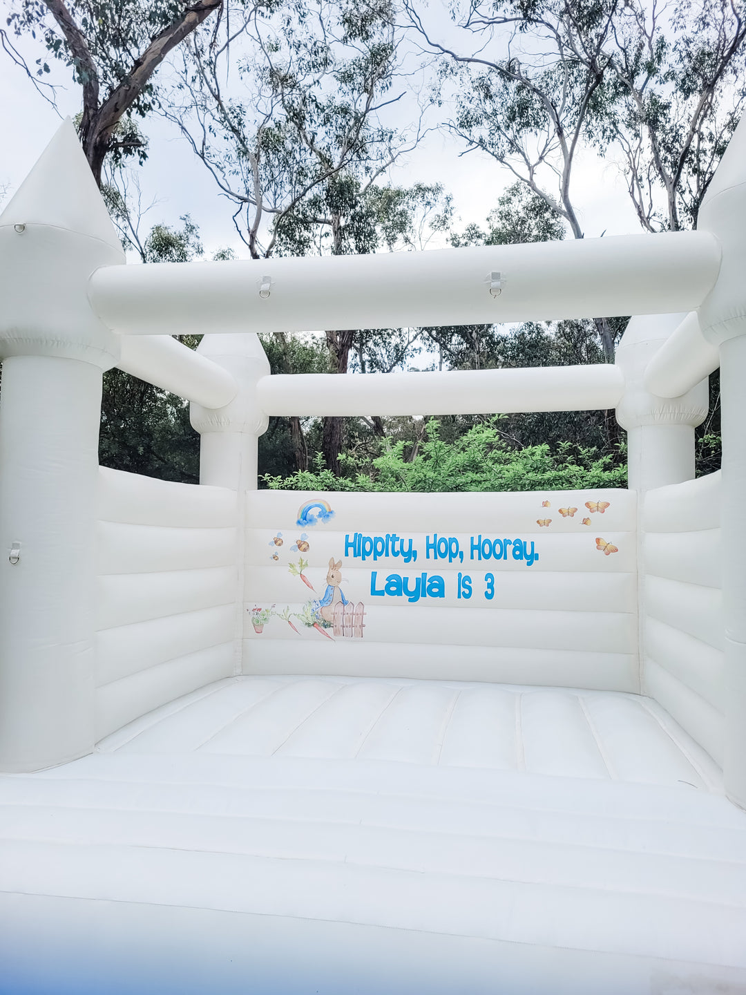 Front view from the step of a white bouncy castle with Peter Rabbit, rainbow and butterfly decals and a Layla is 3 name decal