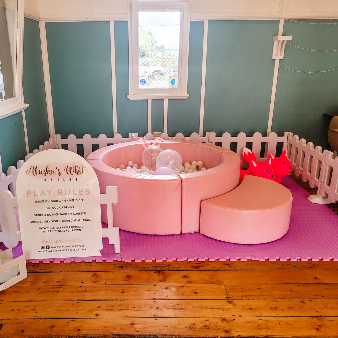 Pink ball pit with white balls, confetti balloons and two hot pink animal hoppers on purple mats beside a play rules sign