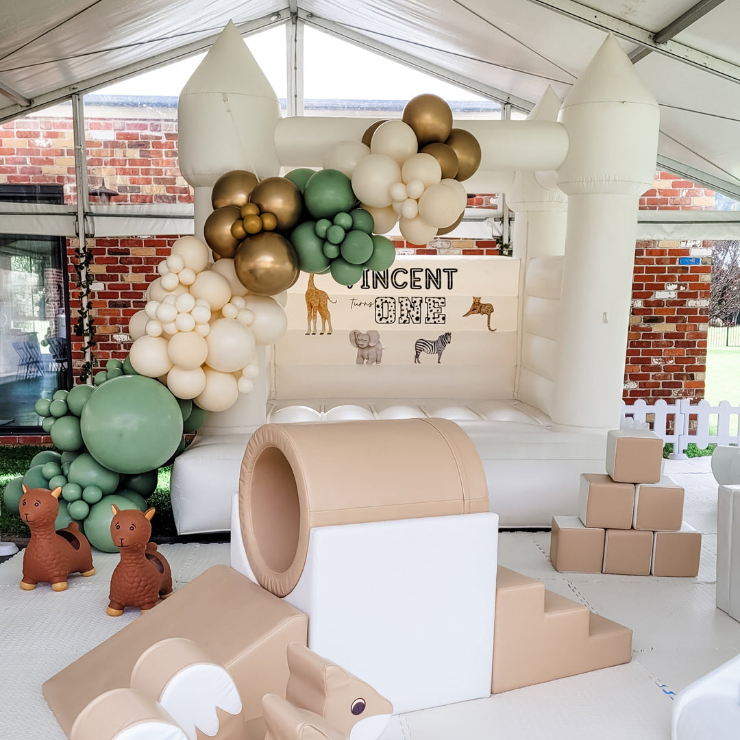 Alex white bouncy castle in a marquee with a sage, cream and gold balloon garland and safari decal reading Vincent turns One