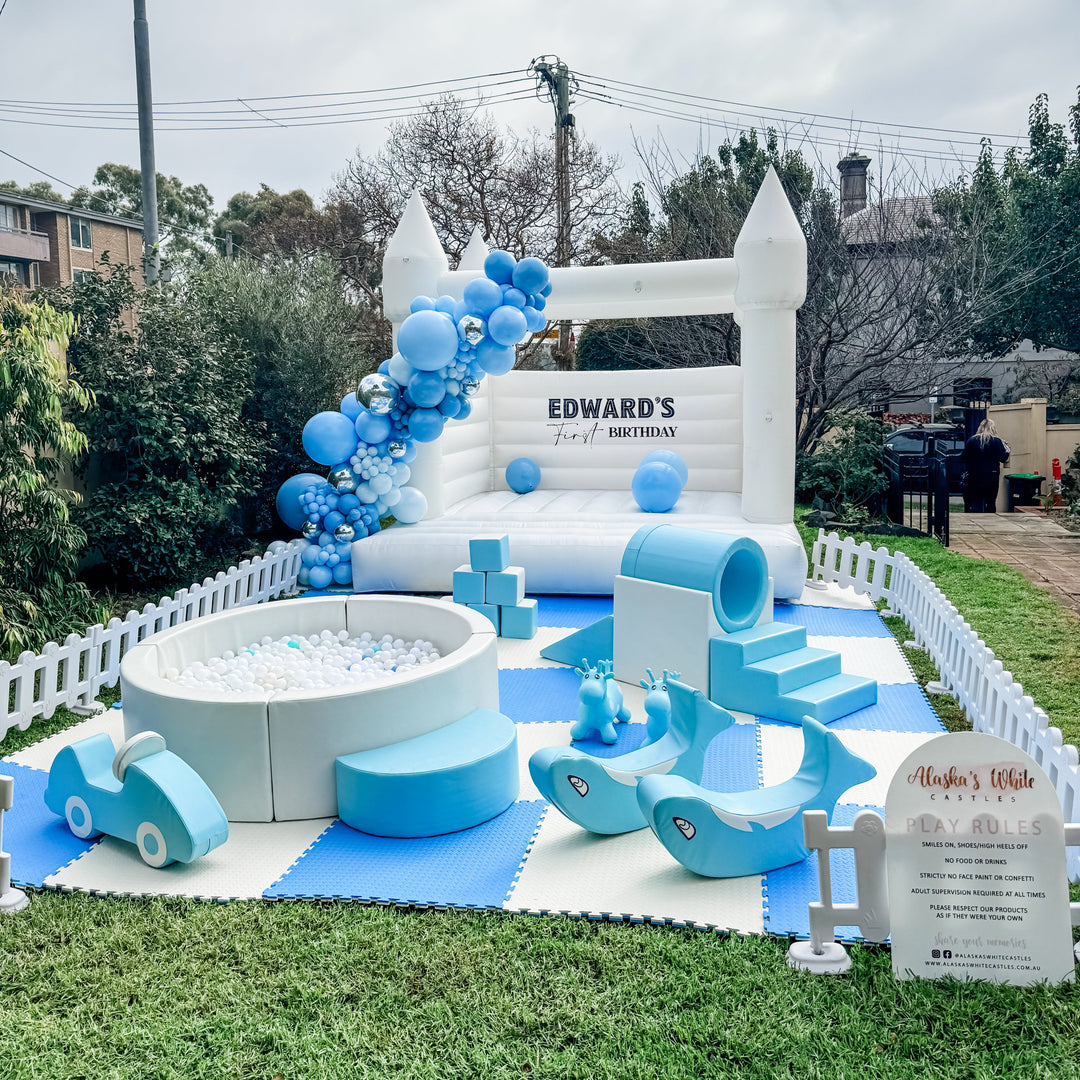 Alaska white bouncy castle with blue balloon garland and Edward's First Birthday decal behind a fenced blue soft play setup