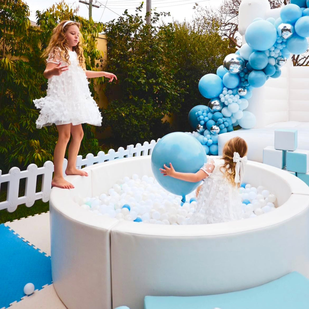 Two children in white dresses at the white round ball pit with pale blue balls, one holding a large blue balloon