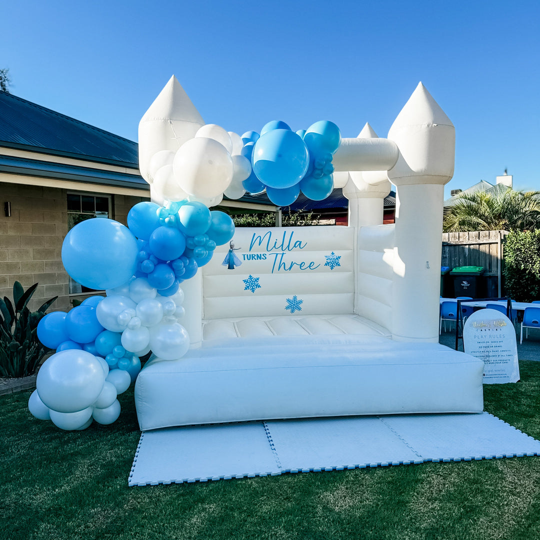 Alex white bouncy castle in a backyard with a blue and white balloon garland and Frozen themed Milla turns Three decal