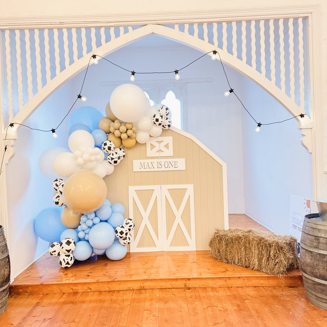 Barn door backdrop with a Max is one sign, blue, tan and cow print balloon garland and a hay bale under festoon lights
