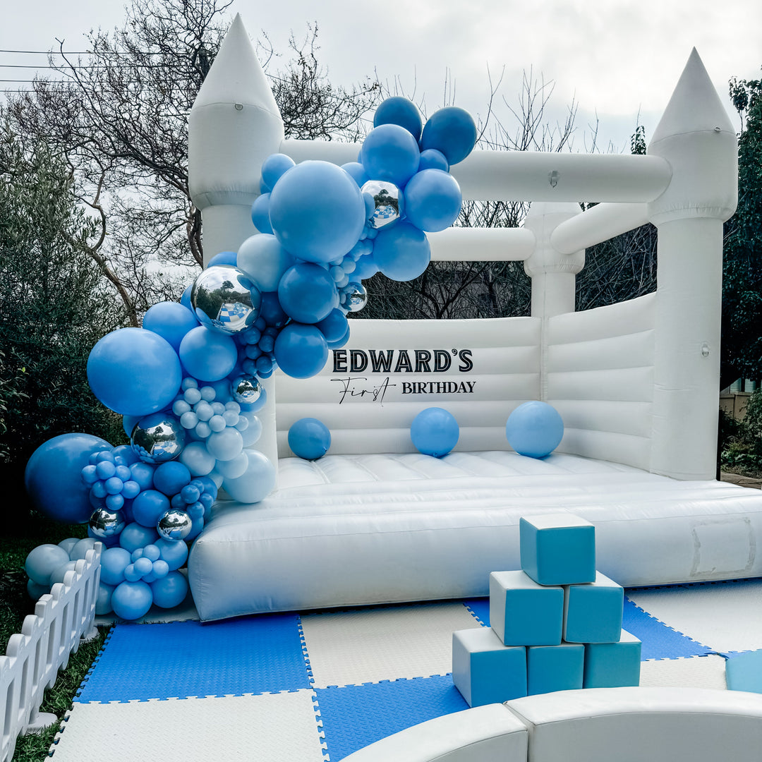 White bouncy castle with a blue balloon garland, stacked blue blocks, a white ball pit and picket fence in a leafy backyard