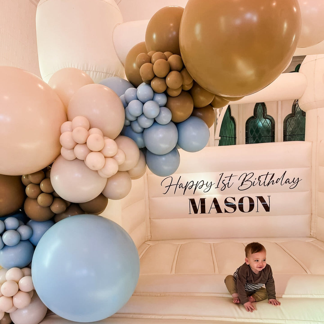 Close-up of the Mason caramel, blush and dusty blue balloon garland with a baby sitting on the bouncy castle floor below