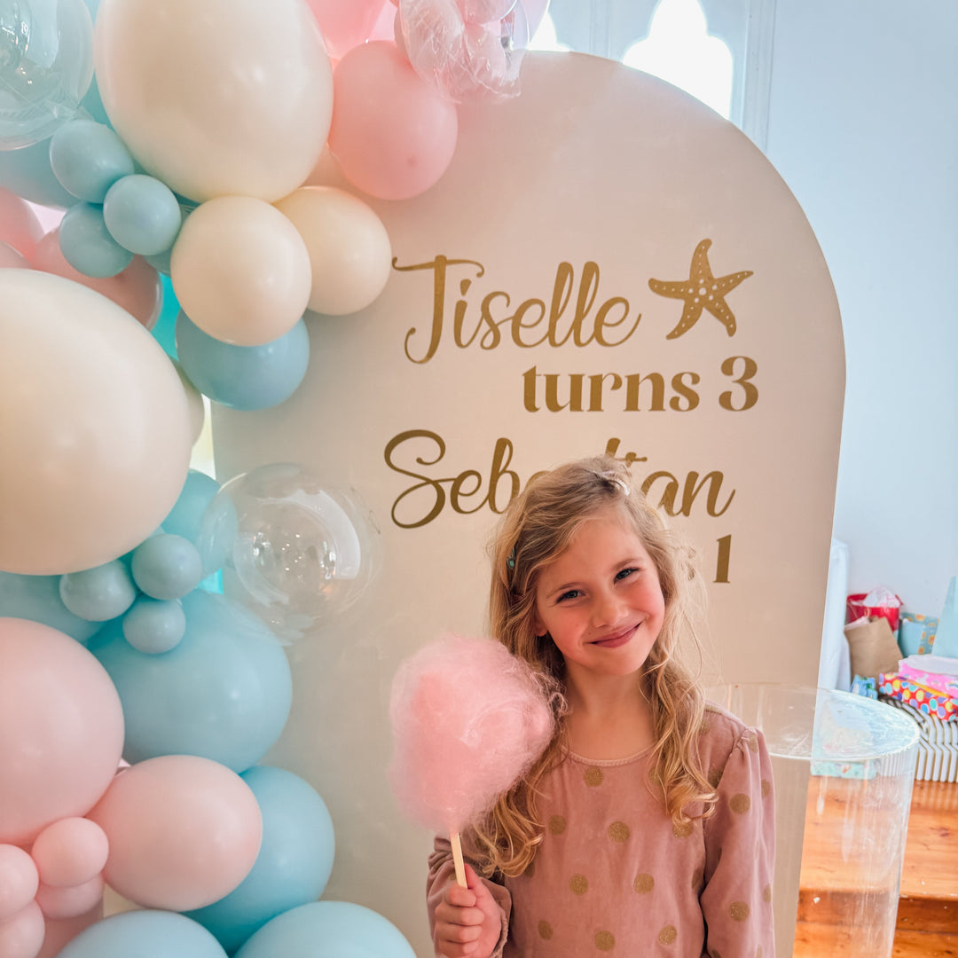 Close-up of a child holding pink fairy floss in front of the cream arch backdrop with a gold Jiselle turns 3 decal
