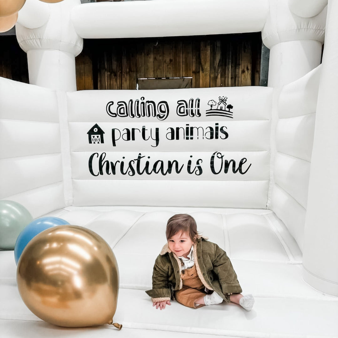 Toddler sitting on the white bouncy castle floor beside a large chrome gold balloon under the Christian is One decal