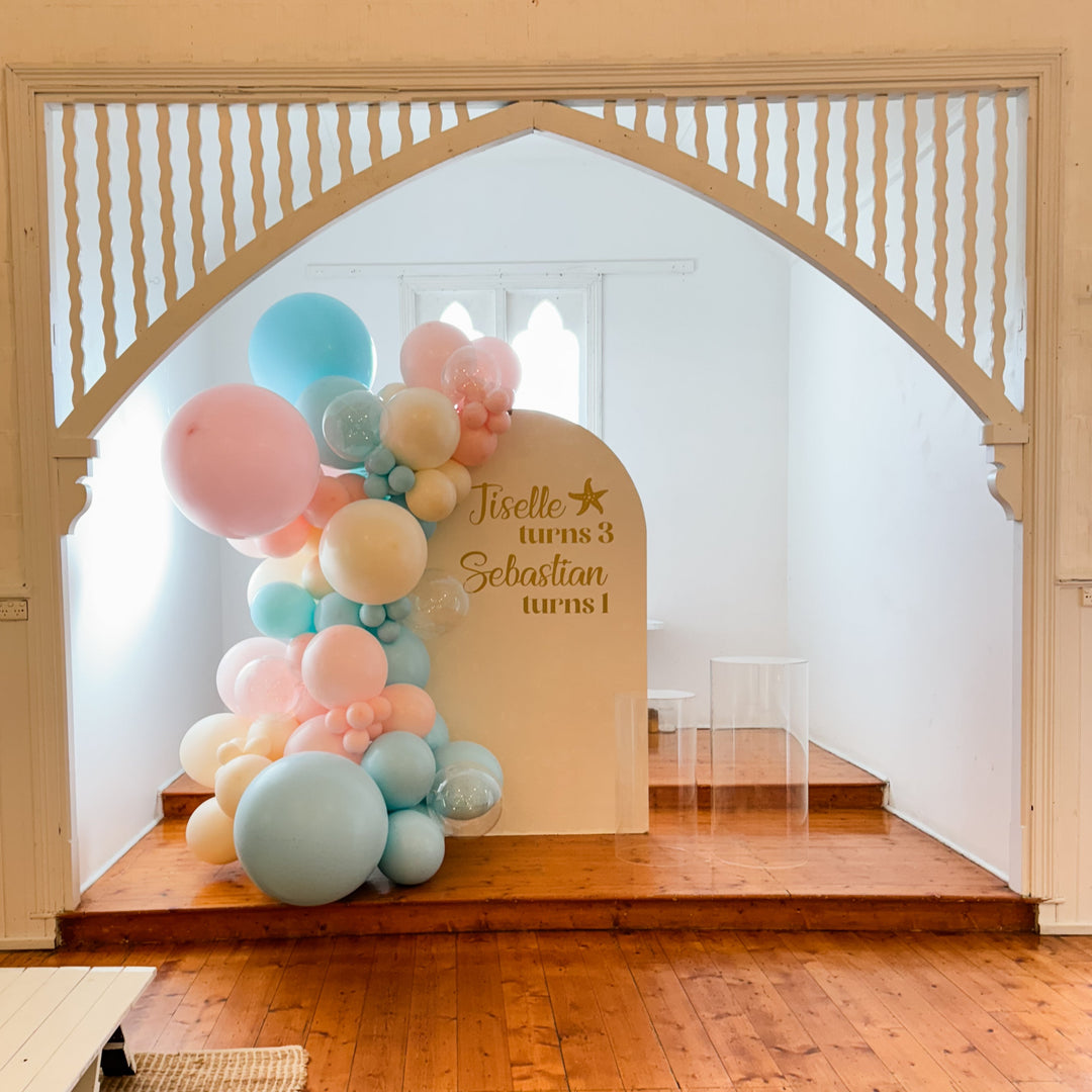 Pastel pink and blue balloon garland on a cream arch backdrop with a gold Jiselle turns 3 Sebastian turns 1 decal on a stage