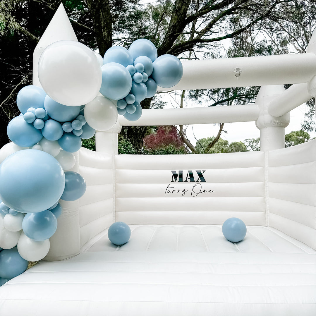 Alaska white bouncy castle front view with a dusty blue and white balloon garland and Max turns One decal under gum trees