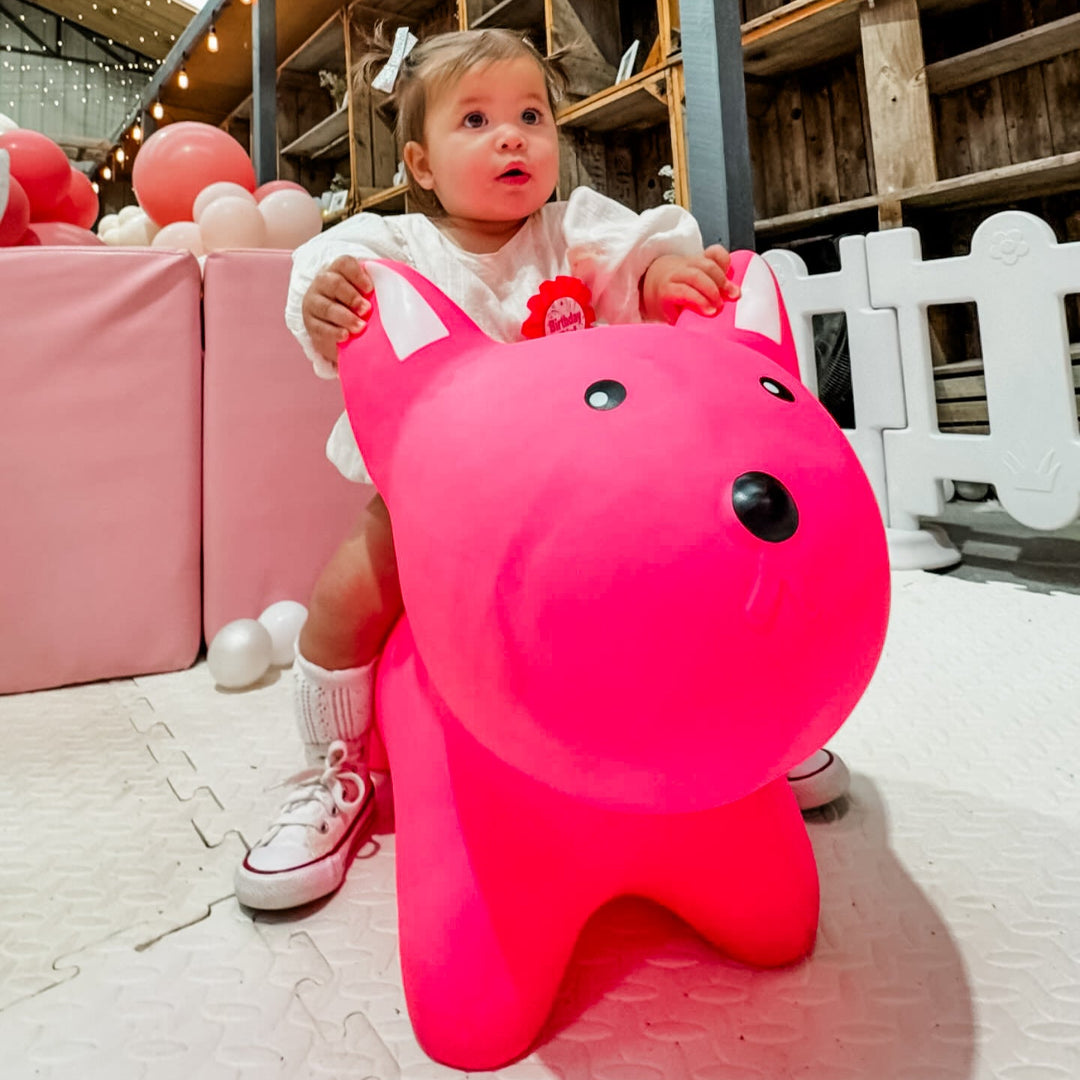Toddler in a white outfit sitting on a hot pink bunny hopper toy on cream foam mats beside pink soft play and balloons