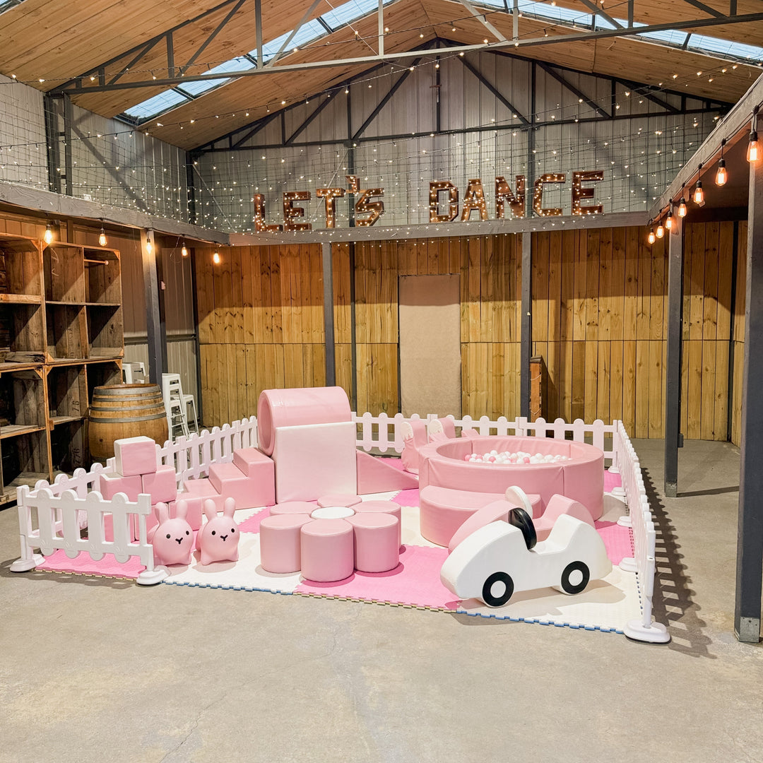 Wide view of the baby pink soft play under a Let's Dance sign and fairy lights in a rustic timber venue, white foam car in front