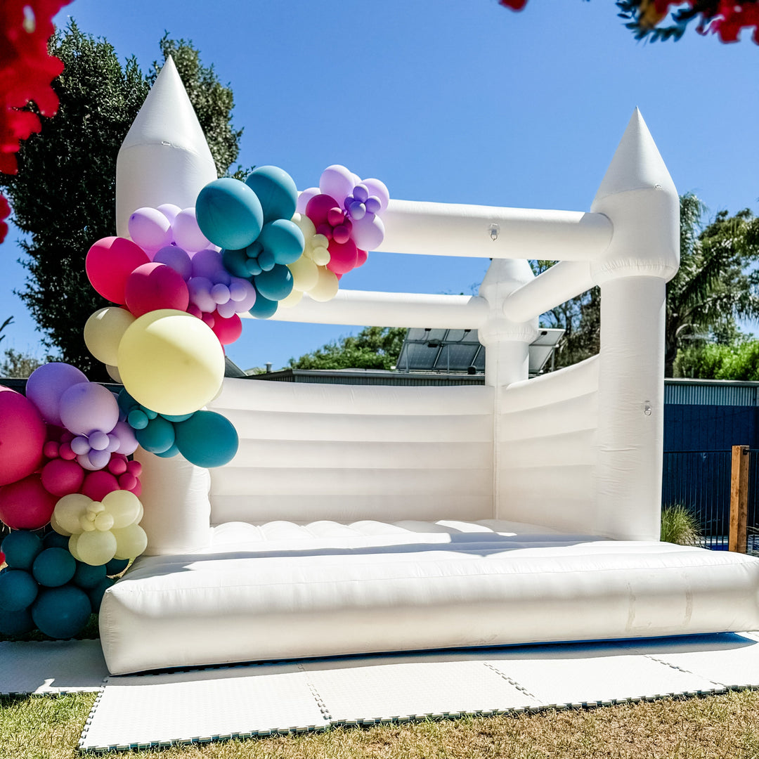 Wider view of the white bouncy castle with a teal, magenta, purple and lemon garland on one tower and foam mats on the grass