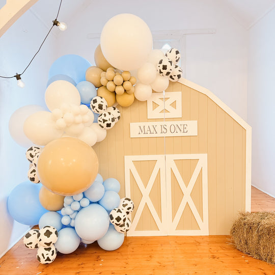 Rodeo balloon garland in blue, tan, white and cow print beside a barn door backdrop with a Max is one sign and a hay bale
