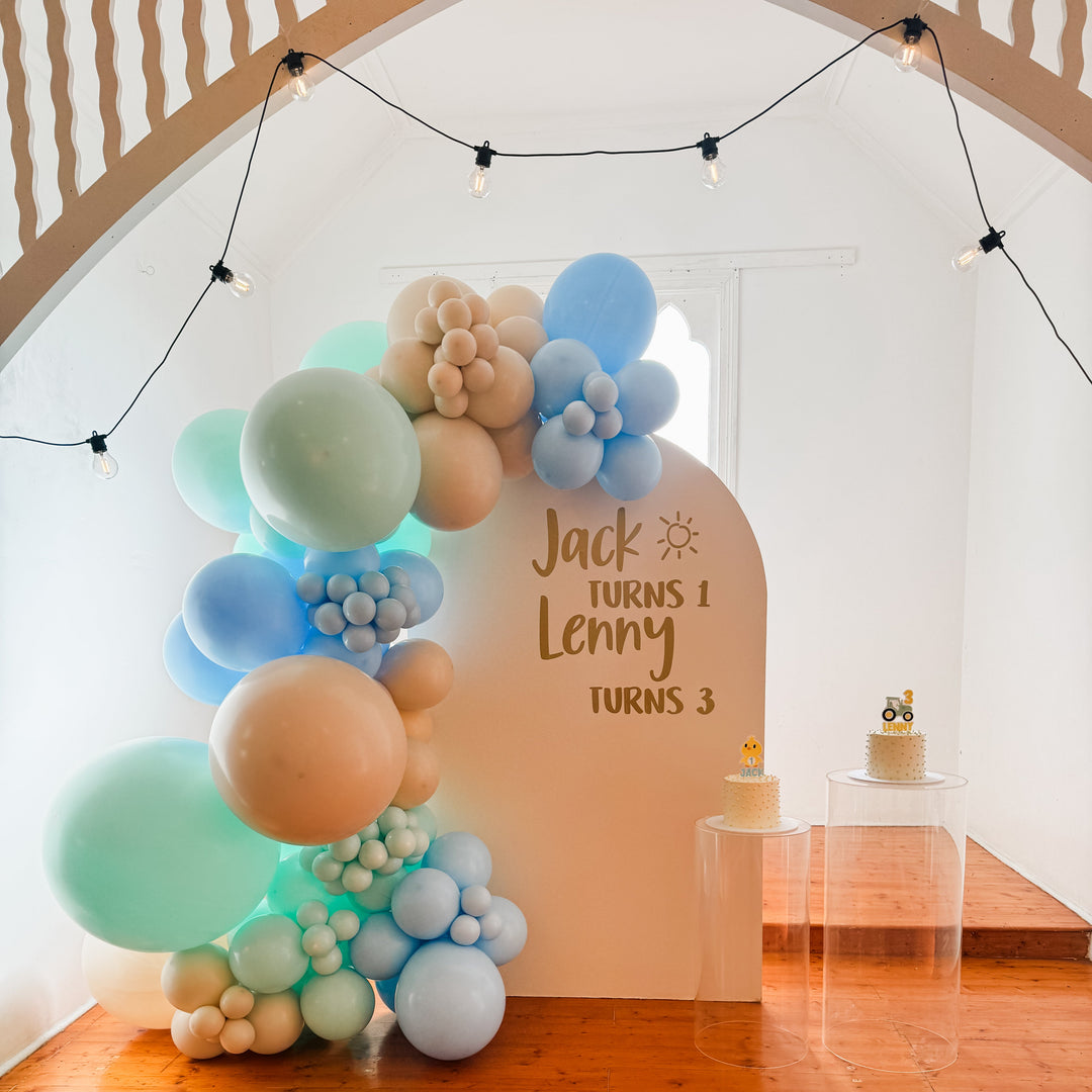 Jack and Lenny balloon garland on a cream arch backdrop with a gold Jack turns 1 Lenny turns 3 decal and two cakes on plinths