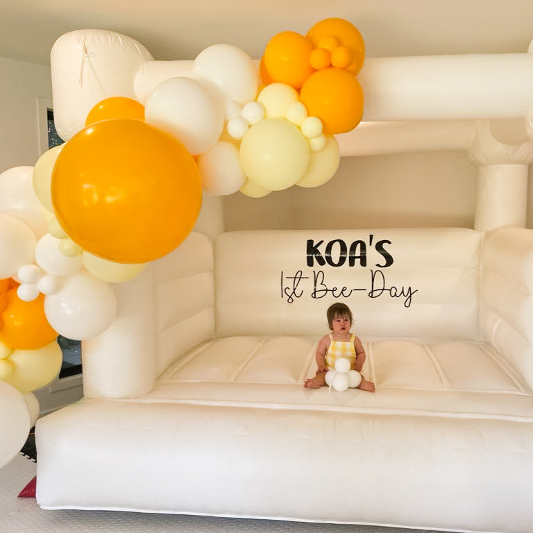 Baby sitting inside the mini white bouncy castle holding white balloons below a Koa's 1st Bee-Day decal and yellow garland