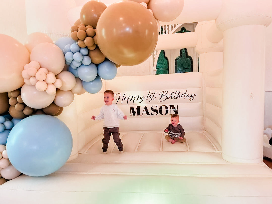 Two children playing inside the white bouncy castle beneath the Mason balloon garland with stained glass windows behind