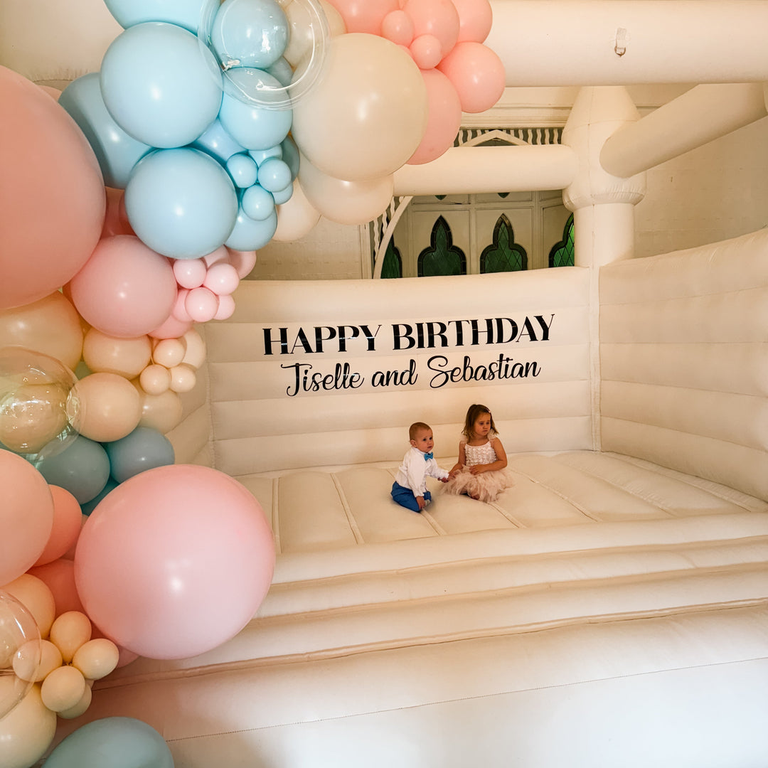 Two children sitting inside a white bouncy castle under a Jiselle and Sebastian birthday decal with the pastel garland above