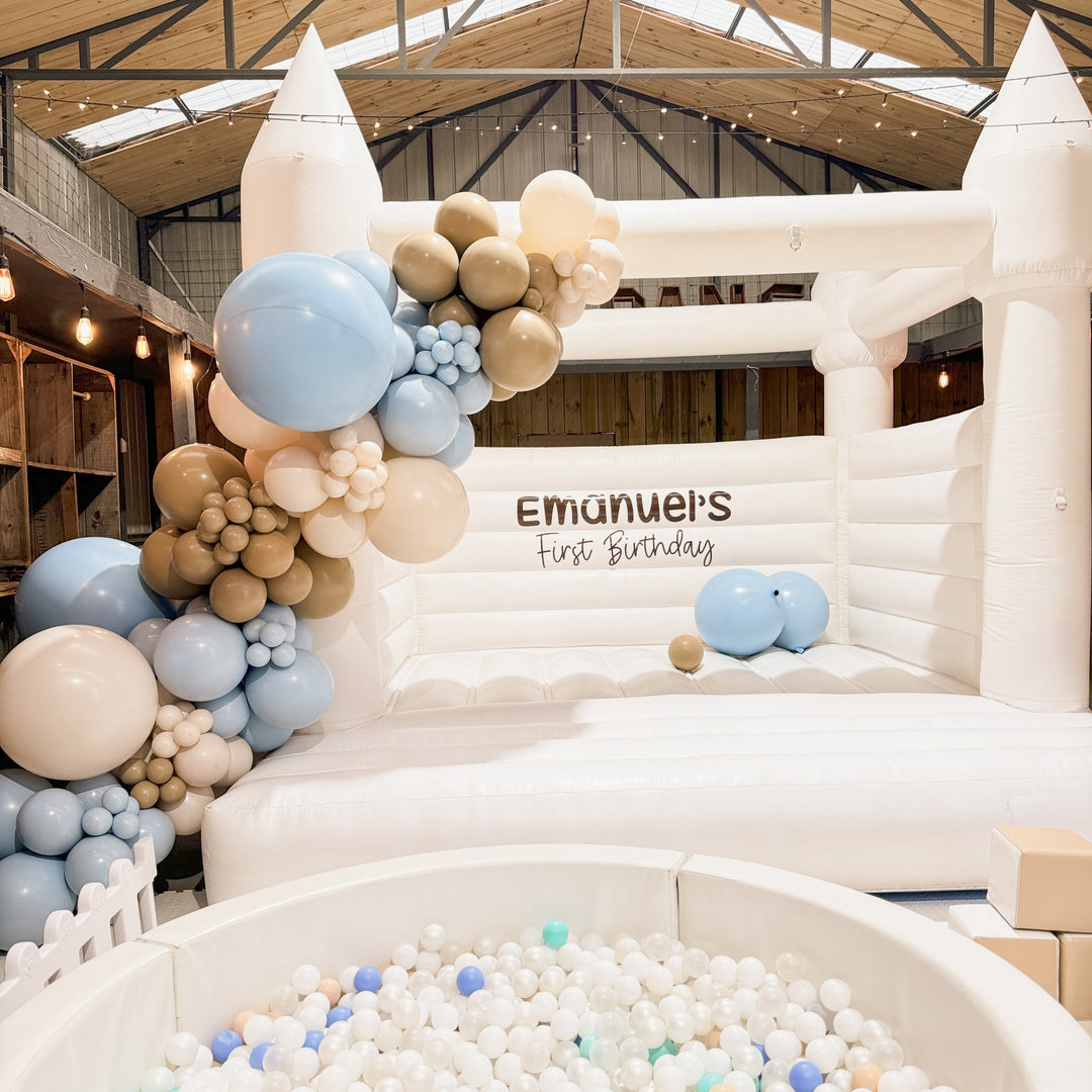 Pale blue, tan and cream balloon garland on a white bouncy castle with an Emanuel's First Birthday decal, ball pit in front