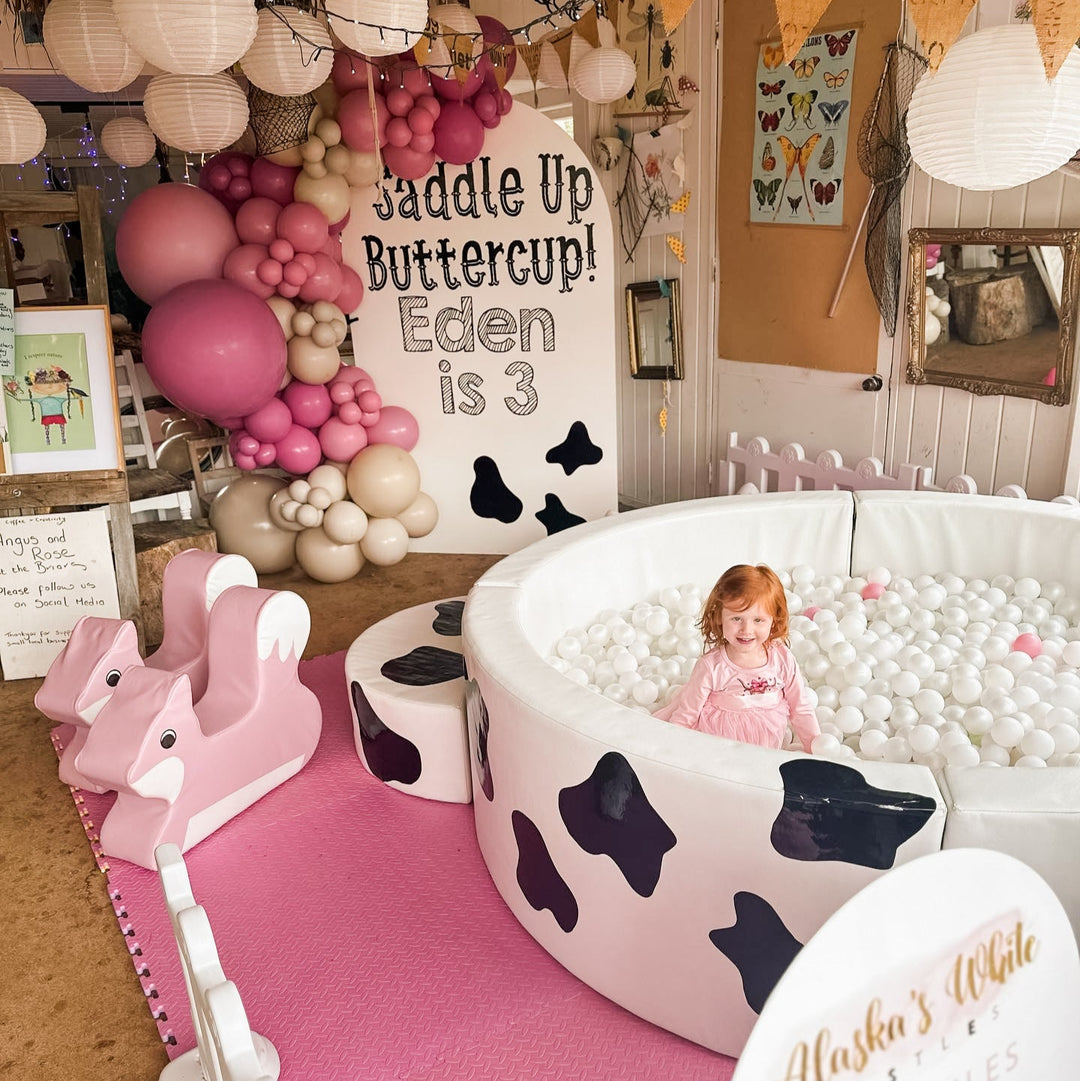 Child sitting among white balls in the cow print Elsa ball pit at a farm party with a Saddle Up Buttercup Eden is 3 sign