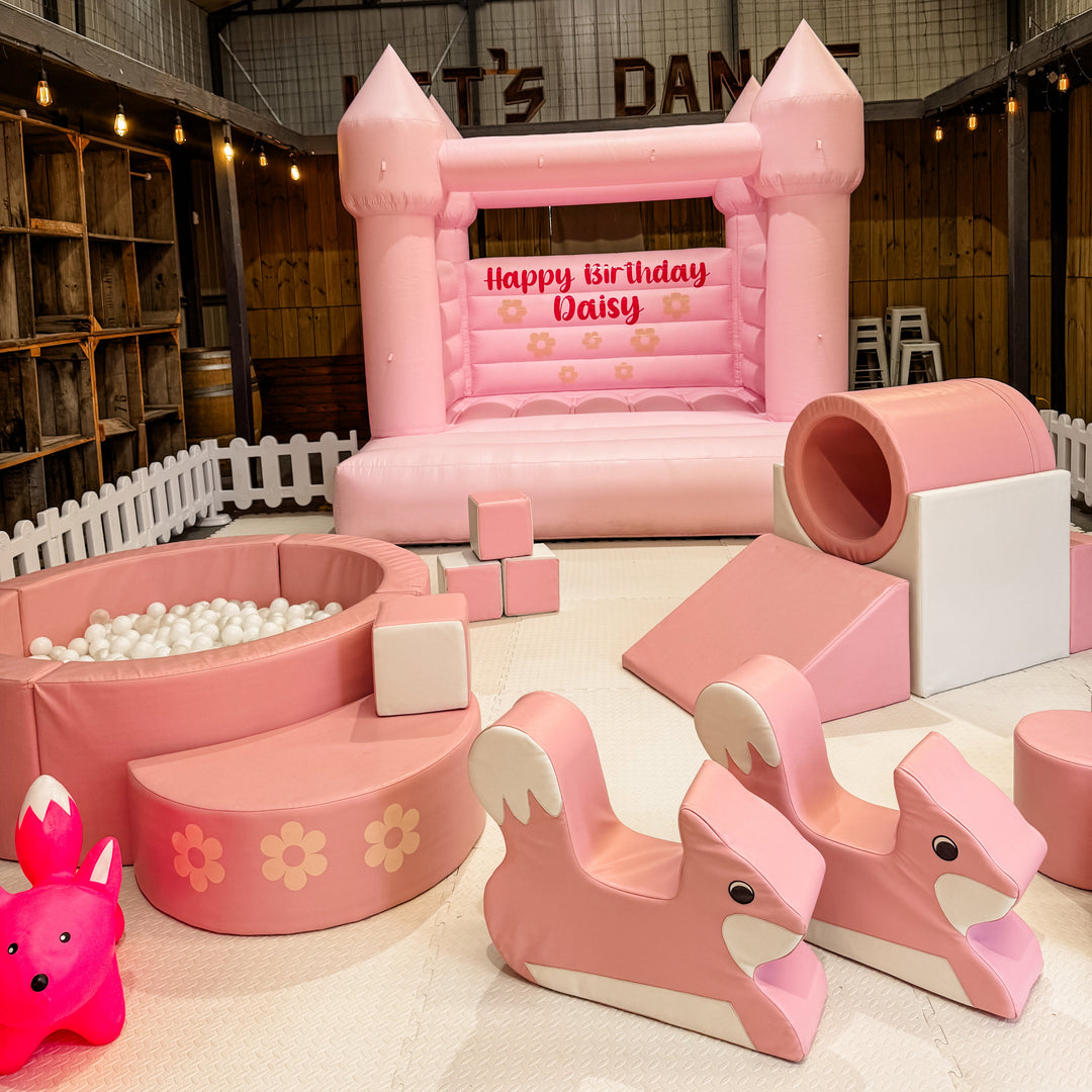 Pink bouncy castle with a Happy Birthday Daisy decal behind dusty pink soft play, a ball pit, rocking animals and a bunny hopper