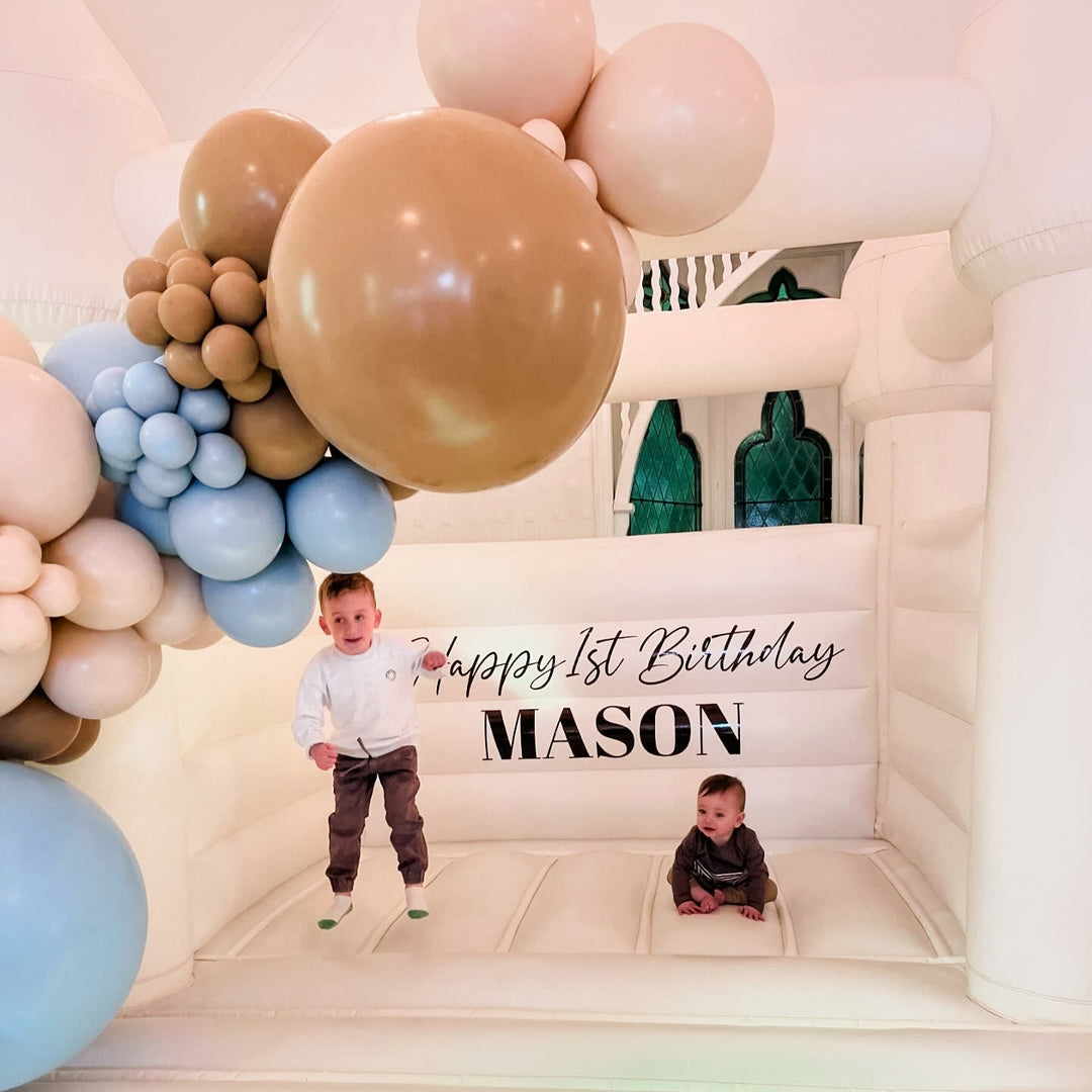 Toddler jumping and a baby sitting inside a white bouncy castle under a Happy 1st Birthday Mason decal and caramel garland