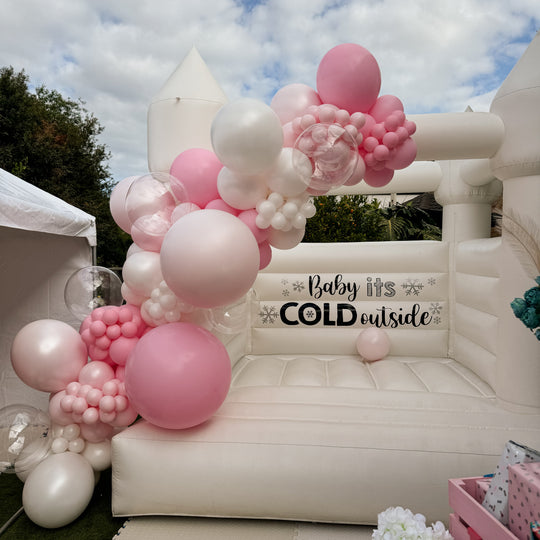 Bella balloon garland in pink, blush and white on a white bouncy castle with a Baby its Cold outside decal for party hire in Melbourne