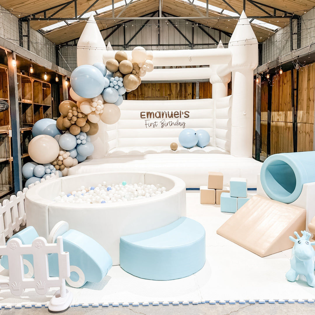 Alaska white bouncy castle with Emanuel's First Birthday decal behind a white ball pit, blue tunnel and soft play blocks