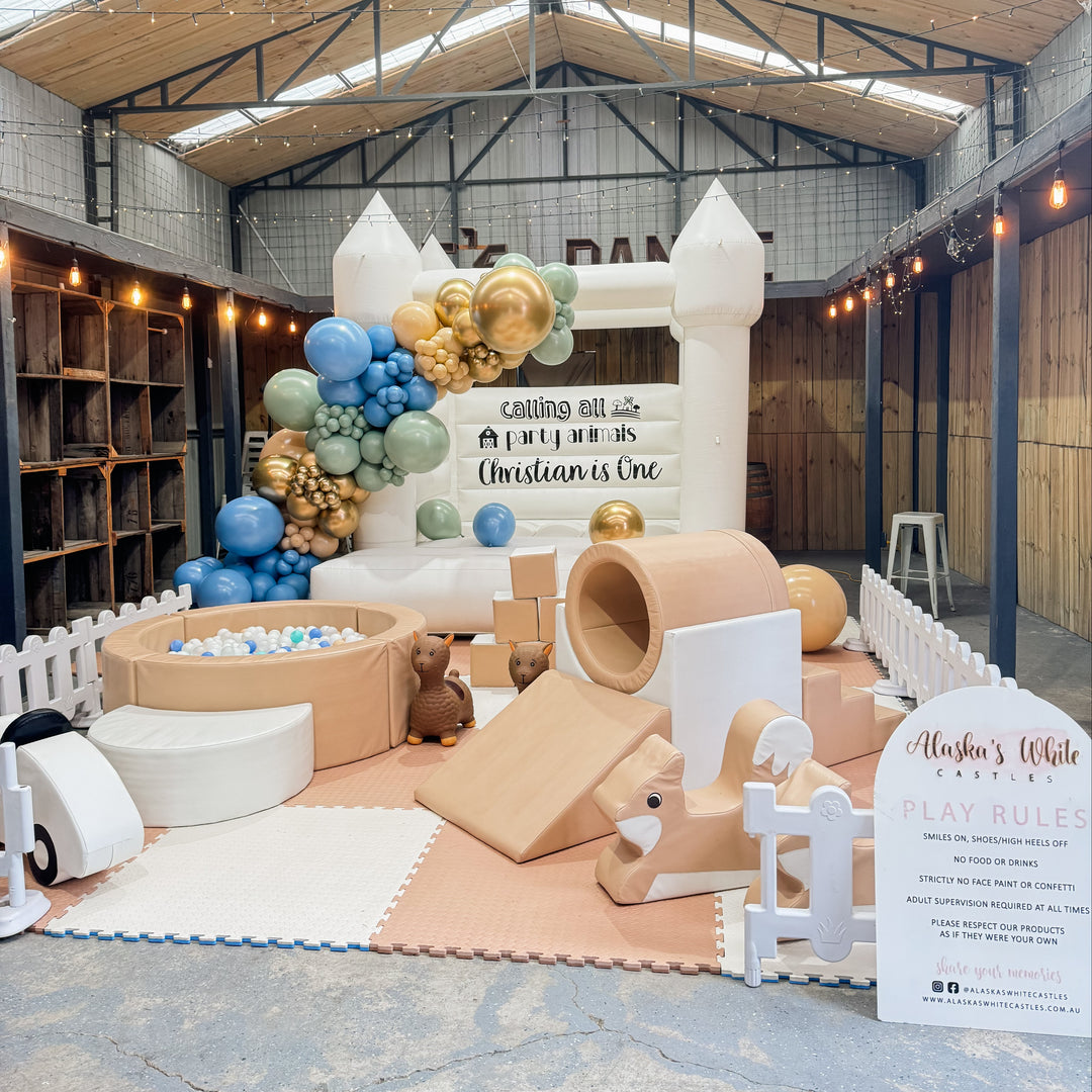 Wide view of a warehouse party with a white bouncy castle, blue and gold balloon garland, tan soft play and play rules sign