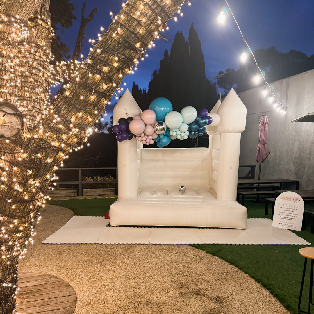 White bouncy castle with a teal, blue, pink and purple balloon garland at night beside a tree wrapped in fairy lights