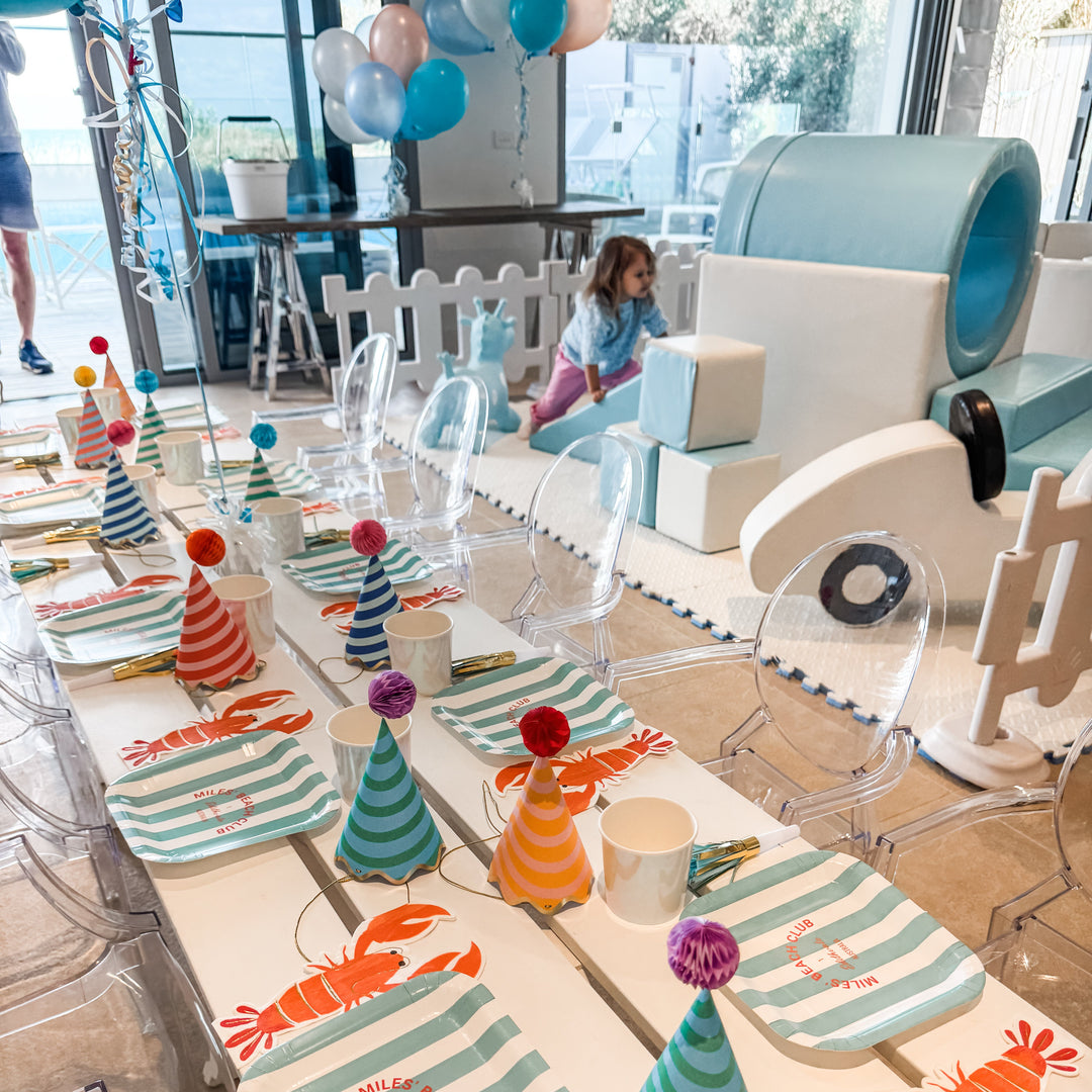 Kids party table with lobster print plates and striped party hats in front of a fenced baby blue soft play setup with a toddler