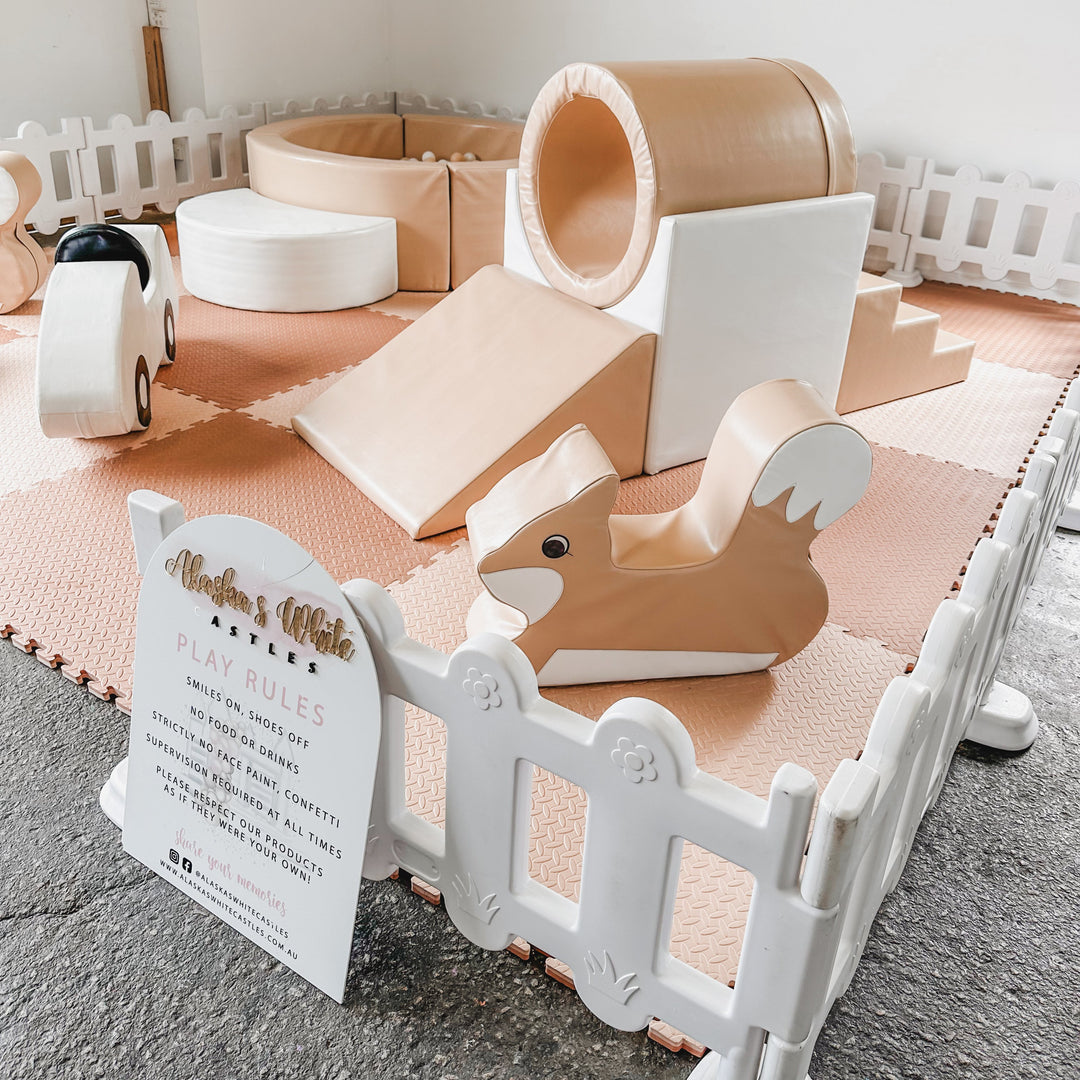Tan fox rocker in front of a tan tunnel slide and ball pit on pink mats, with a play rules sign and white picket fence