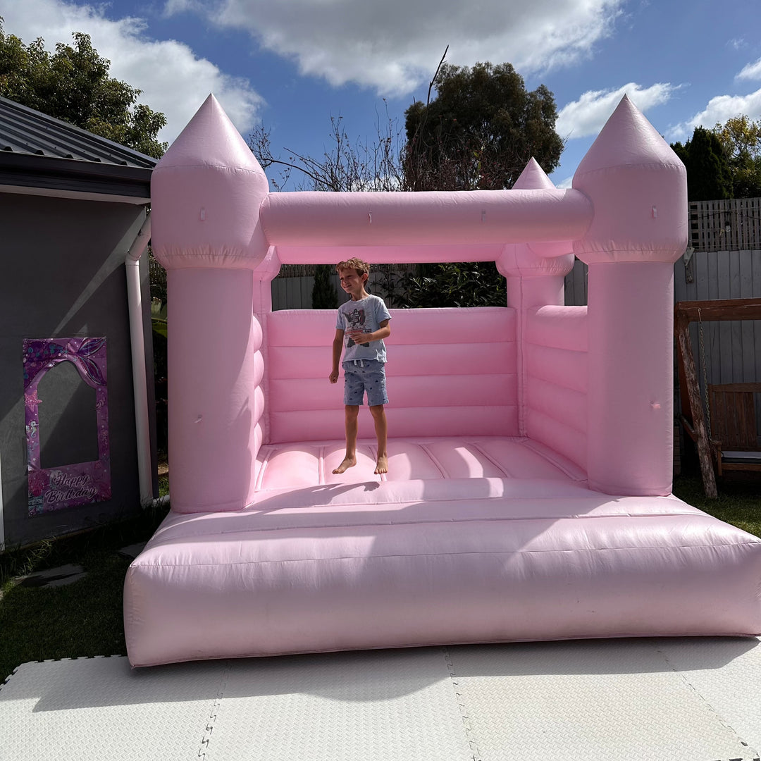 Pink bouncy castle set up on cream foam mats in a backyard with a child bouncing inside and a timber fence behind