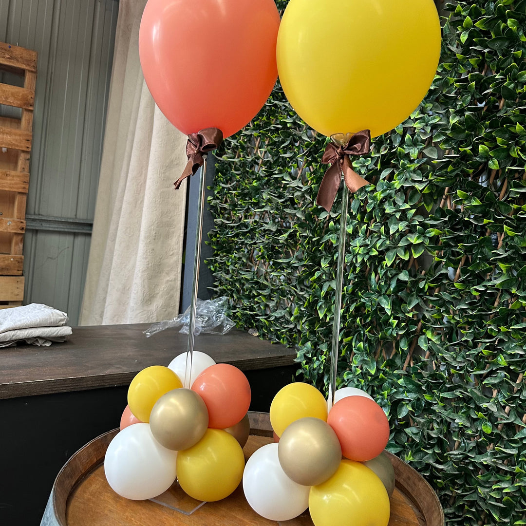 Close-up of two acrylic tabletop balloon stands on a wine barrel with coral and yellow balloons and brown ribbon bows