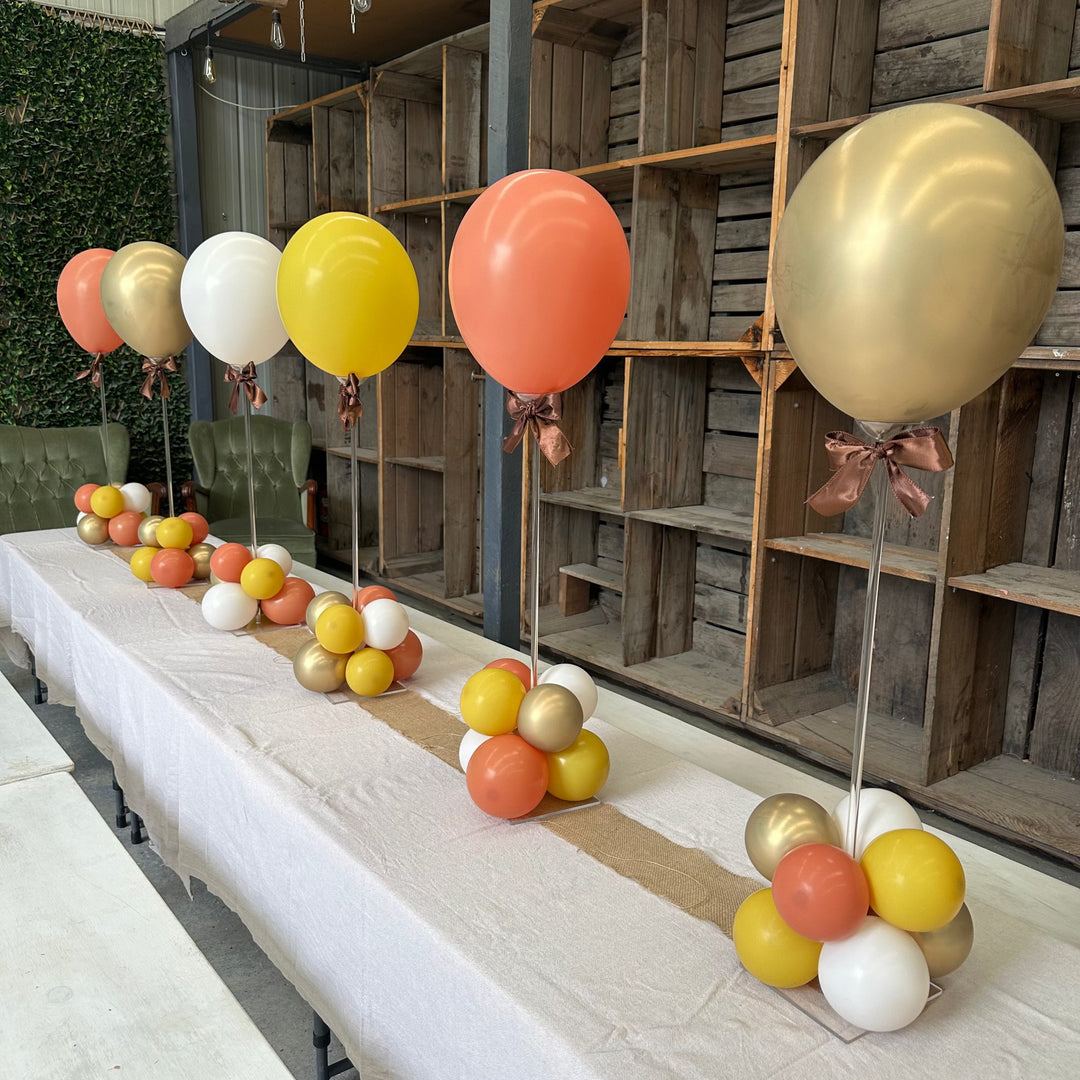 Row of five acrylic tabletop balloon stands along a long white table with coral, yellow, white and gold balloons