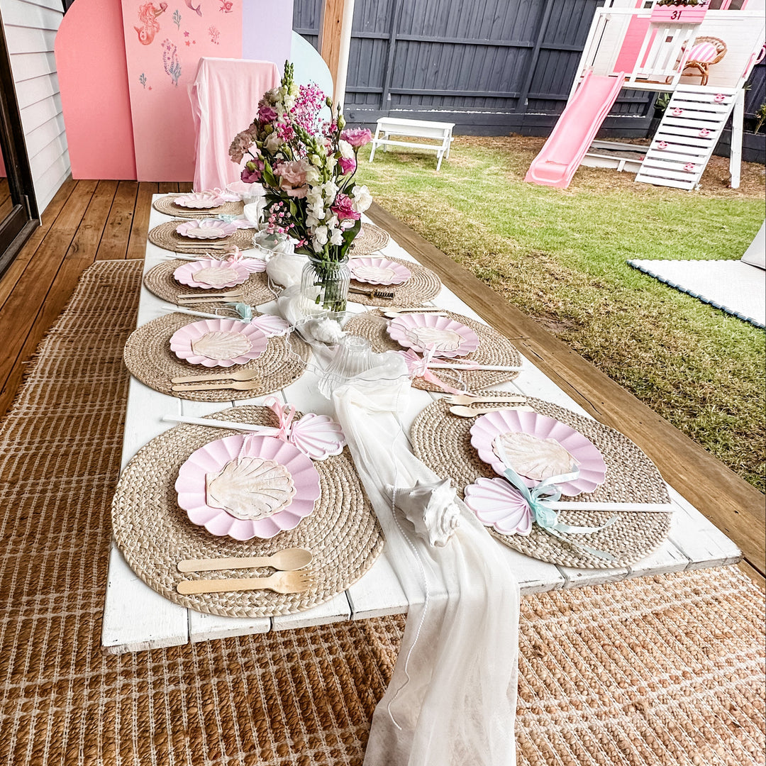 Low white trestle table on a jute rug styled for a mermaid party with pink shell plates and rattan placemats on a deck