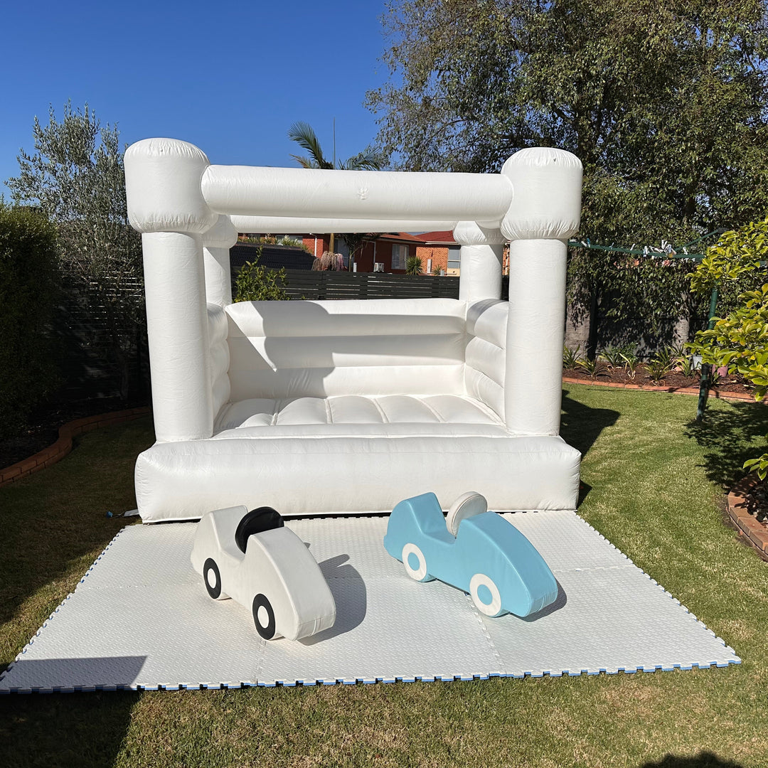 Mini white bouncy castle set up plain on a backyard lawn with a white and a blue soft play ride-on car on white foam mats