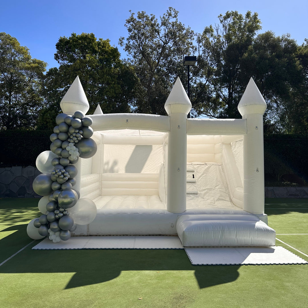 William white bouncy castle with slide and a silver, grey and clear balloon garland set up on a tennis court on a sunny day