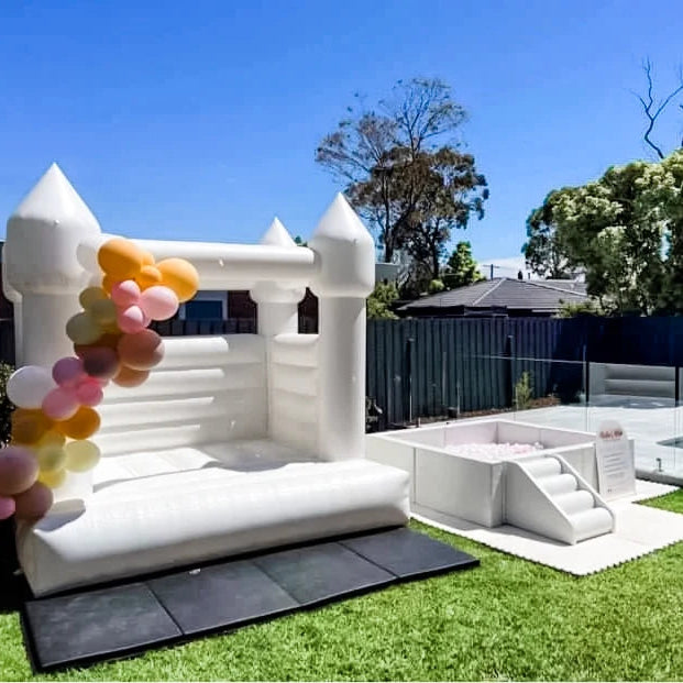 White bouncy castle with an orange, mustard and pink balloon garland on a sunny lawn, ball pit and steps beside a pool fence