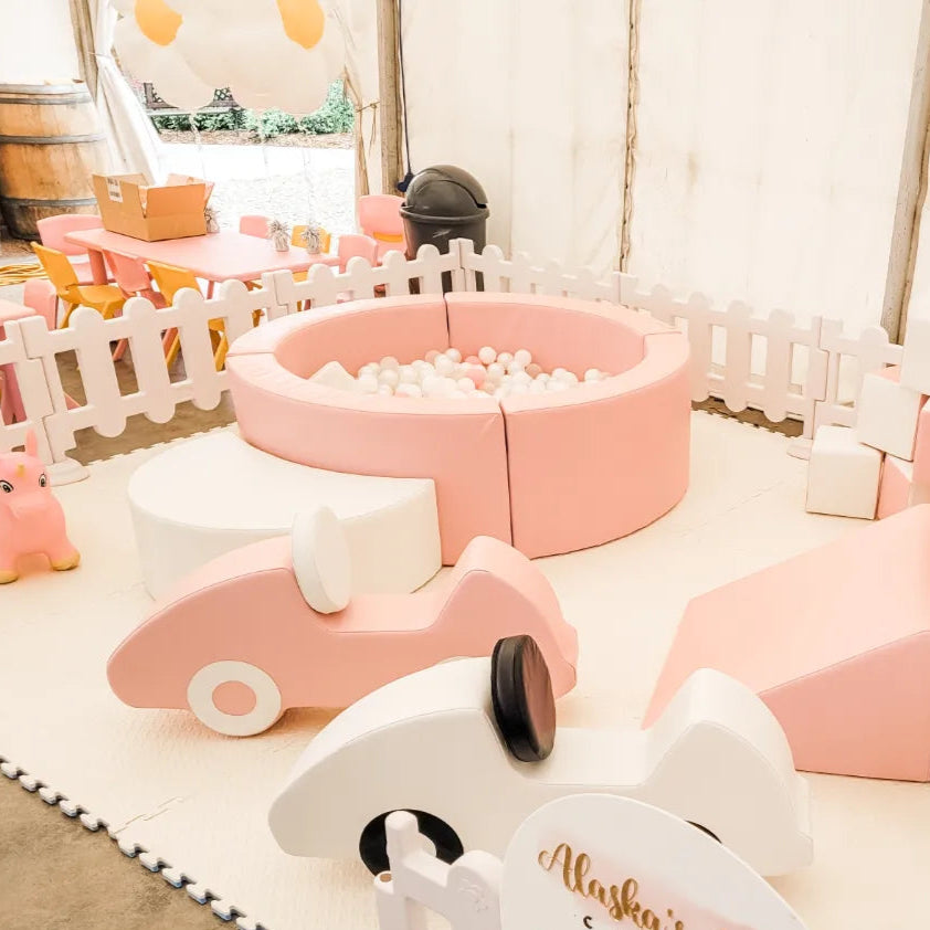Pink round ball pit with white balls inside a white picket fence in a marquee with pink soft play cars and a kids party table