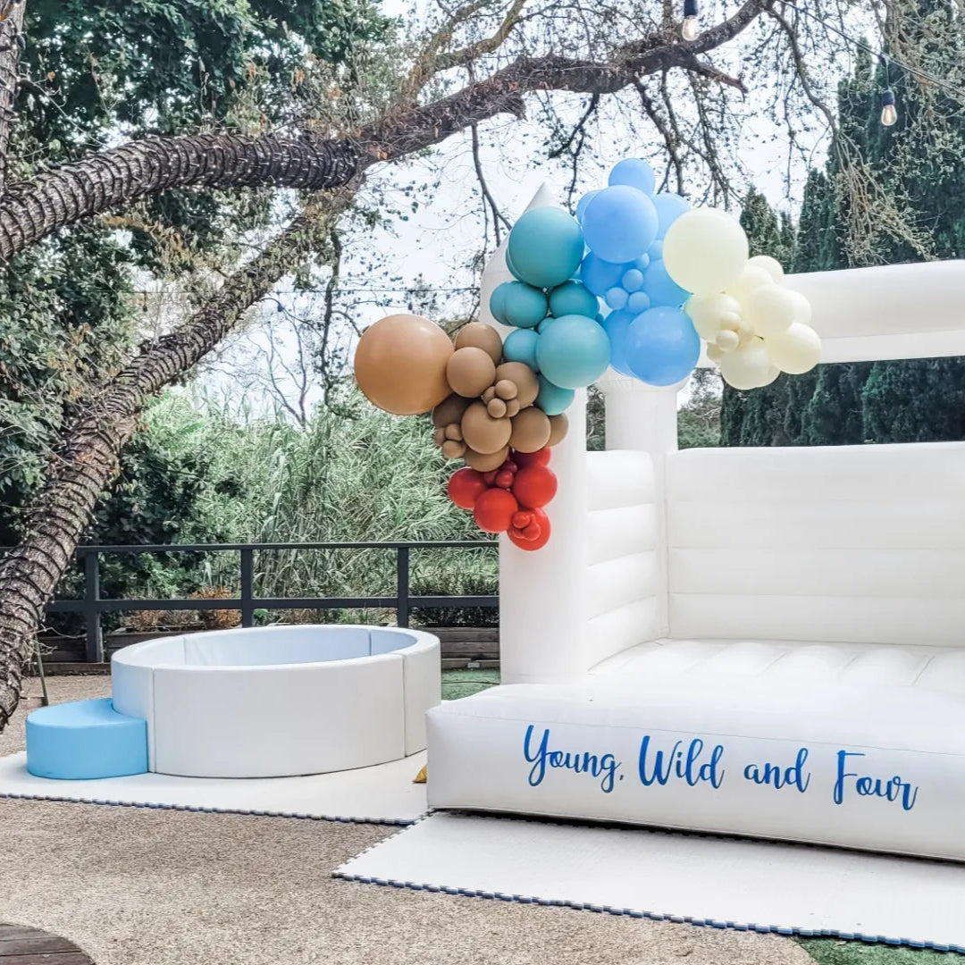 Elsa white round ball pit with a blue step beside a white bouncy castle with a Young, Wild and Four decal and balloon garland