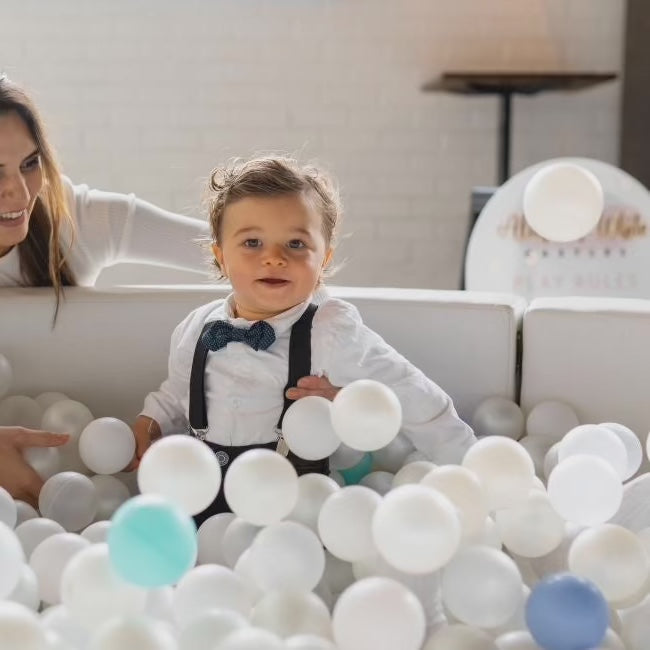 Toddler in a bow tie sitting in a white ball pit of white, clear and mint balls as an adult leans in by the play rules sign